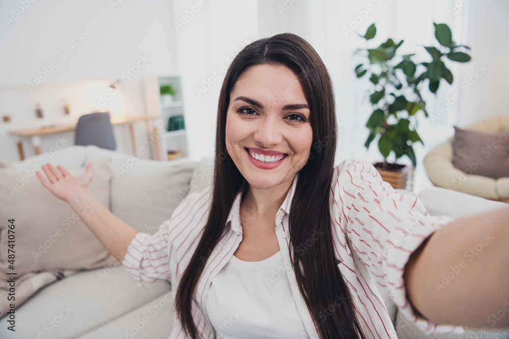 Self-portrait of attractive cheerful hospitable long-haired woman sitting on divan inviting home indoors Self-portrait of attractive cheerful hospitable long-haired woman sitting on divan inviting home indoors