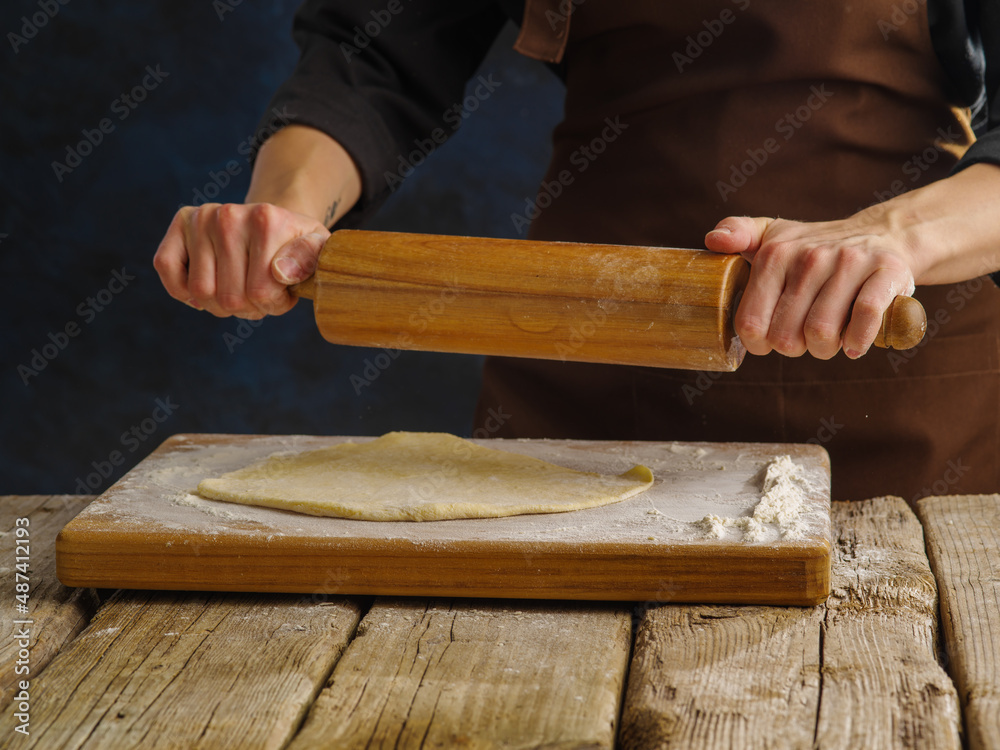 Preparation of dough products by a professional chef in a professional kitchen. The chef rolls out the dough on a wooden cutting board with a rolling pin. Bakery, restaurant, hotel, cafe, pastry shop Preparation of dough products by a professional chef in a professional kitchen. The chef rolls out the dough on a wooden cutting board with a rolling pin. Bakery, restaurant, hotel, cafe, pastry shop