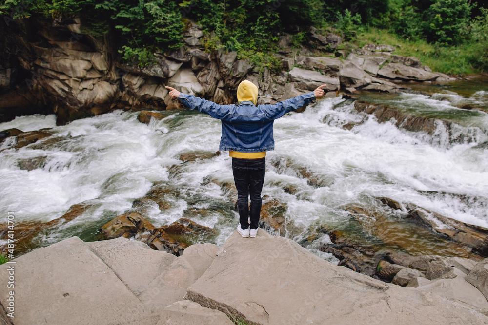 Happy male tourist stands with his back with raised arms in the fresh air in the forest by the mountain river. Happy male tourist stands with his back with raised arms in the fresh air in the forest by the mountain river.