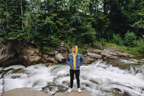 Happy male tourist standing outdoors in the forest near the mountain river.