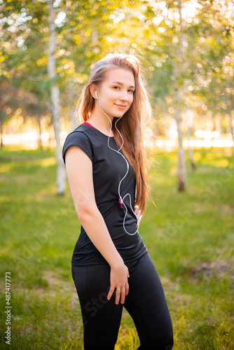 Portrait of a beautiful smiling girl in a sports uniform with flowing hair listening to music in headphones on the background of the park. © Михаил Гута