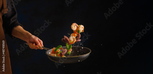 Fototapeta Naklejka Na Ścianę i Meble -  On a dark background, a frying pan in the chef's hand with seafood. Levitation. Seafood recipe book. Advertising, banner. There is an empty space for the recipe.