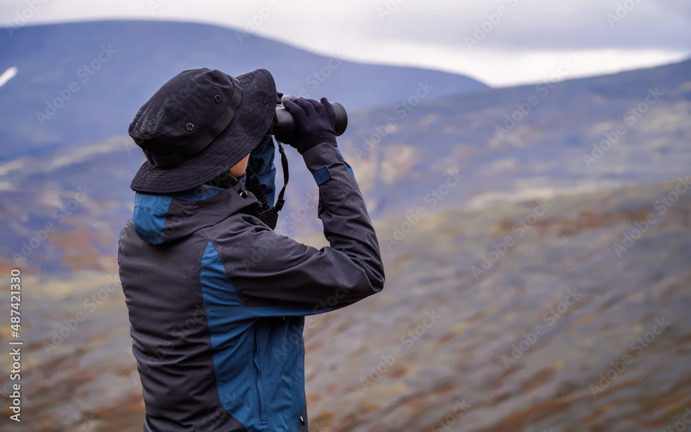 A woman in a hat on a mountain slope looks through binoculars in autumn against the backdrop of a lake and mountains. view from the back.
