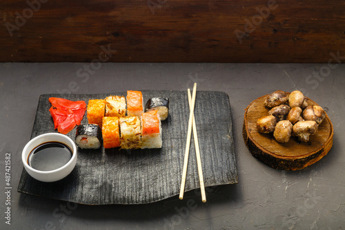 Assorted rolls on a black handmade board next to sticks, soy sauce and baked mushrooms on a dark background.