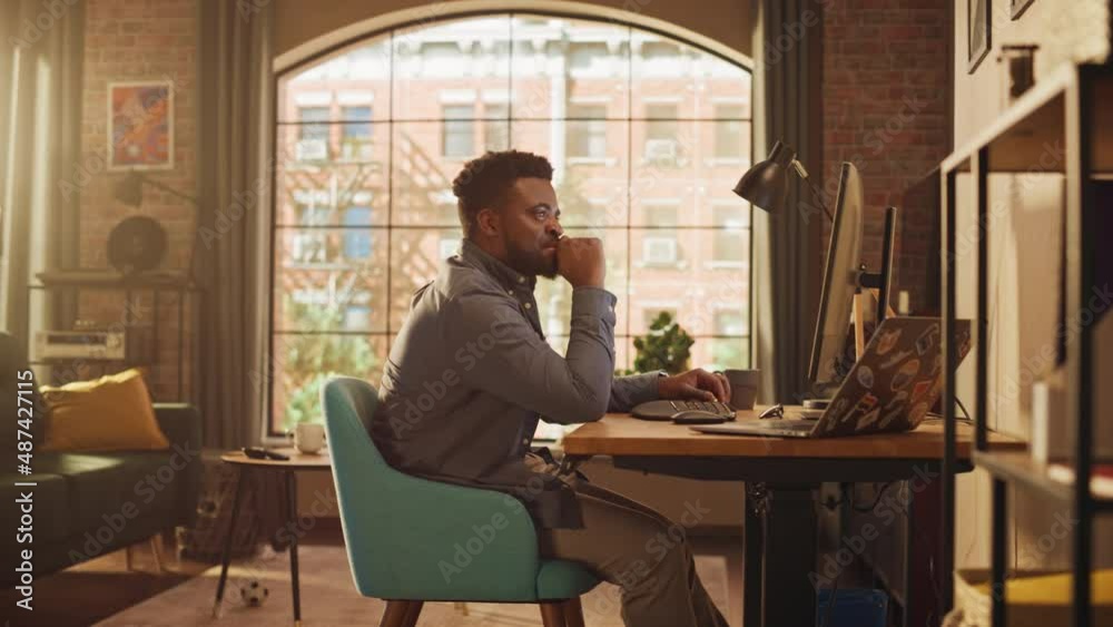 Young Handsome Black Man Working from Home on Desktop Computer in Sunny Stylish Loft Apartment. Creative Male Checking Social Media, Browsing Internet. Urban City View from Big Window.