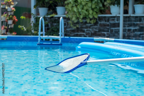 Net-skimmer in a clear water pool with an inflatable mattress. Backyard with flowers in the background. Round frame pool and leaf netting skimmer for cleaning leaves from the surface of the water.
