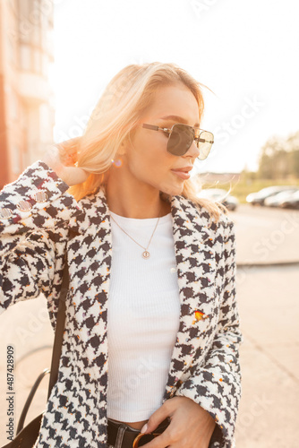 Beautiful young girl blonde with glasses in fashionable casual clothes with a stylish coat, white T-shirt and bag walking on the street at sunset