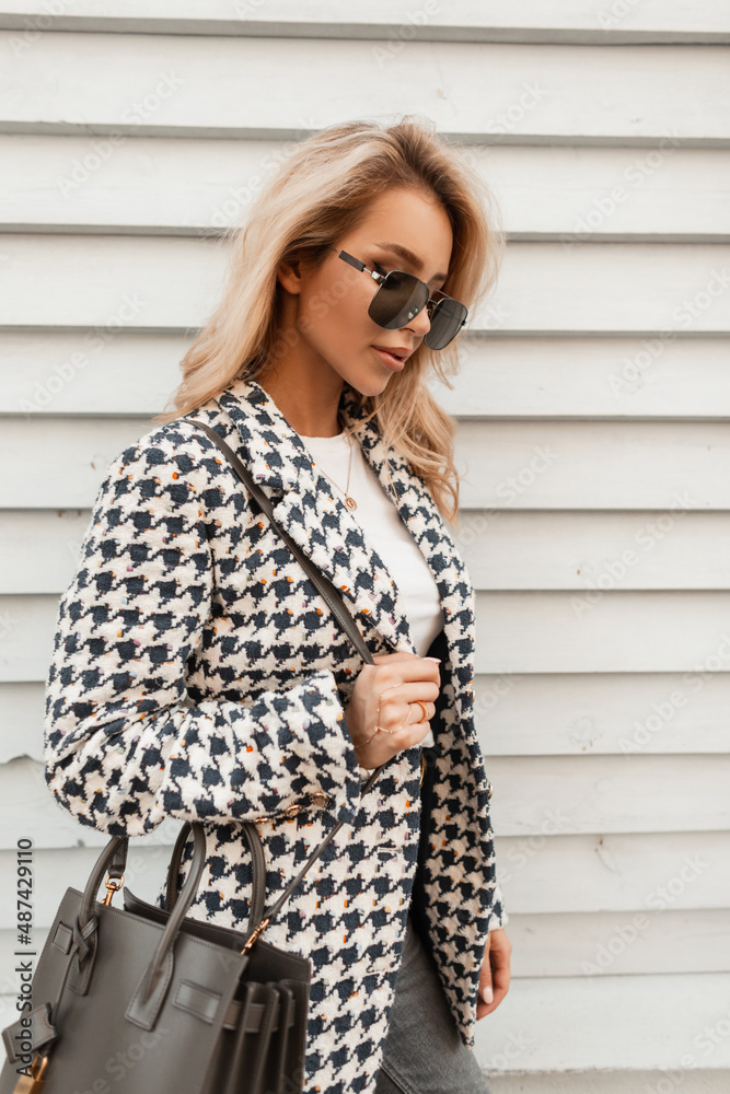 Trendy young blonde hipster woman with sunglasses in fashion outwear with jacket and bag stands near wooden wall Trendy young blonde hipster woman with sunglasses in fashion outwear with jacket and bag stands near wooden wall