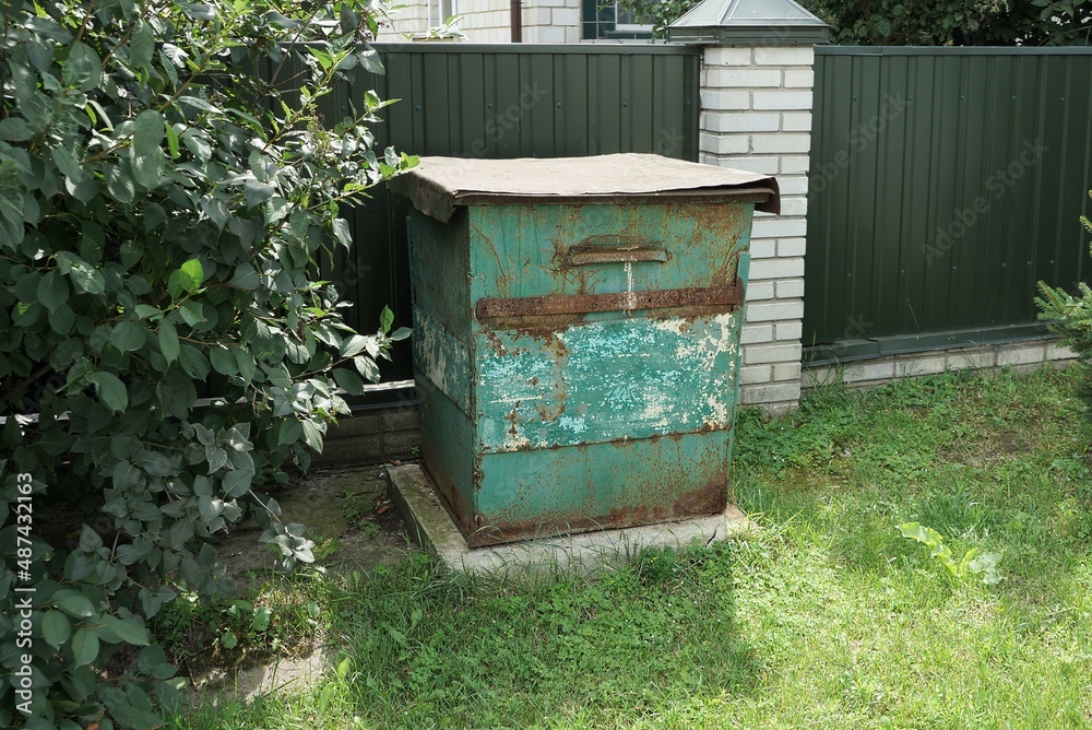 one old rusty closed metal trash can stands in green grass and vegetation on a rural street near an iron fence wall one old rusty closed metal trash can stands in green grass and vegetation on a rural street near an iron fence wall
