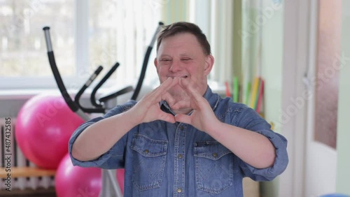 A young man with Down syndrome smiles in love, showing a heart symbol and a shape with his hands. Romantic concept.
