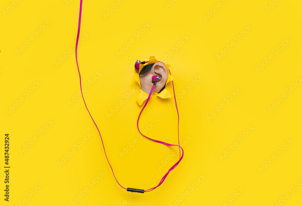 Female ear with pink vacuum headphones in a torn hole in yellow paper. The concept of a music lover, listener of music and audio relaxation. Background with copy space. Female ear with pink vacuum headphones in a torn hole in yellow paper. The concept of a music lover, listener of music and audio relaxation. Background with copy space.