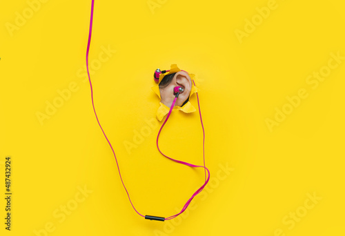 Female ear with pink vacuum headphones in a torn hole in yellow paper. The concept of a music lover  listener of music and audio relaxation. Background with copy space.