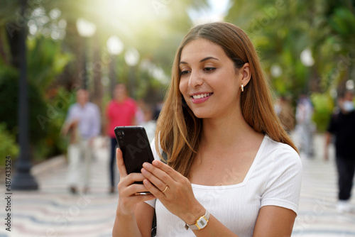Excited young woman watching her smartphone when walking in the street with blurred people on the background. Beautiful girl using mobile app outdoors. Teenager lifestyle technology concept. photo