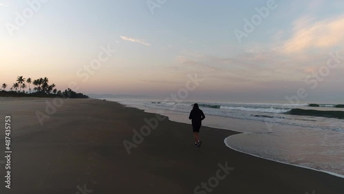 A man is busy with a morning jog at a slow pace along the seashore on the beach next to the ocean at dawn. Sports training, running along the surf line. A man in shorts, hoodies and headphones runs. photo