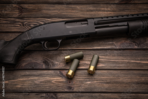 A black 12-gauge pump-action shotgun and ammo are on a wooden table. A smooth-bore weapon with a plastic stock. Dark brown back