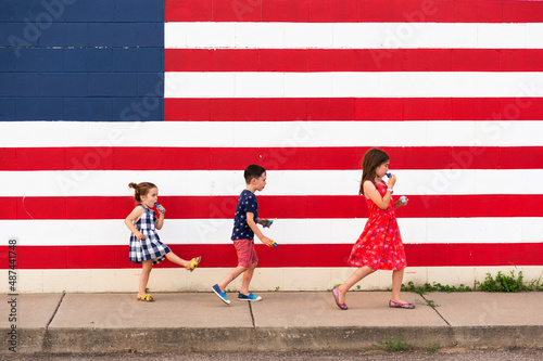 3 kids walk on a sidewalk in front of a large American  flag