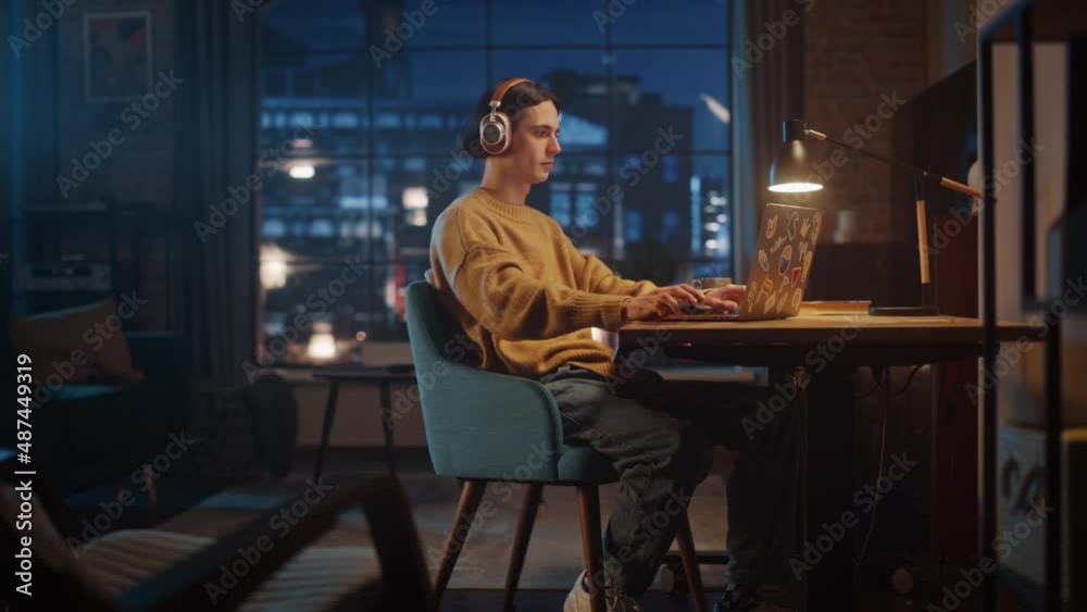 Young Handsome Man Working from Home on Laptop Computer in Stylish Loft Apartment in the Evening. Creative Person Checking Social Media, Browsing Internet. Urban City View from Big Window. Young Handsome Man Working from Home on Laptop Computer in Stylish Loft Apartment in the Evening. Creative Person Checking Social Media, Browsing Internet. Urban City View from Big Window.