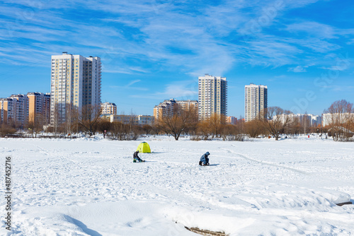 Winter ice fishing, lake, frosty day. A fisherman is engaged in ice fishing in the pond of the city park. Against the background of a multi-storey, modern, residential development.