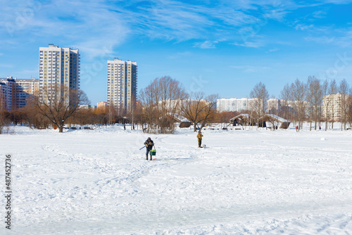 Winter ice fishing, lake, frosty day. A fisherman is engaged in ice fishing in the pond of the city park. Against the background of a multi-storey, modern, residential development.