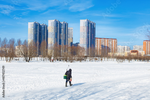 Winter ice fishing, lake, frosty day. A fisherman is engaged in ice fishing in the pond of the city park. Against the background of a multi-storey, modern, residential development.