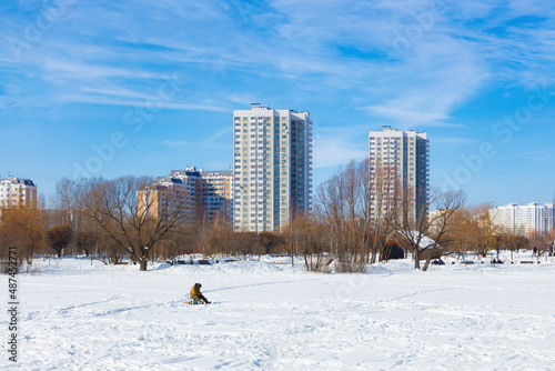 Winter ice fishing, lake, frosty day. A fisherman is engaged in ice fishing in the pond of the city park. Against the background of a multi-storey, modern, residential development.