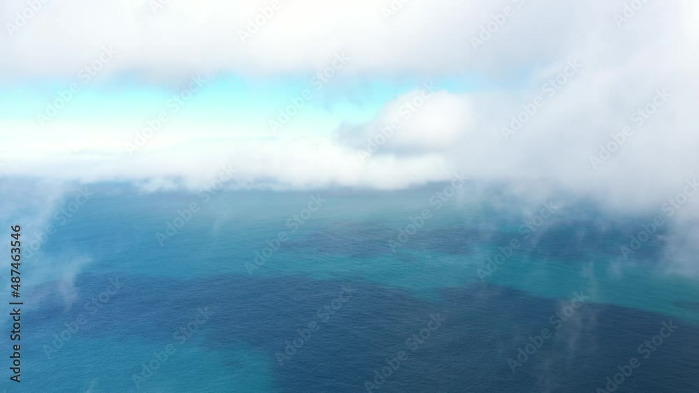 Drone shot flying backwards, surrounded by thin dancing clouds in the blue and turquoise sky with the colorful ocean below with reefs next to Espigao Amorelo mountain in Madeira. Drone shot flying backwards, surrounded by thin dancing clouds in the blue and turquoise sky with the colorful ocean below with reefs next to Espigao Amorelo mountain in Madeira.
