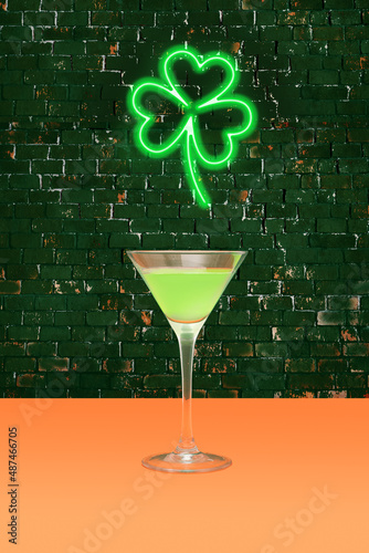 Lucky pub minimal concept. Martini glass with green cocktail on terracotta table. Shamrock green neon sign above on dark brick background.
