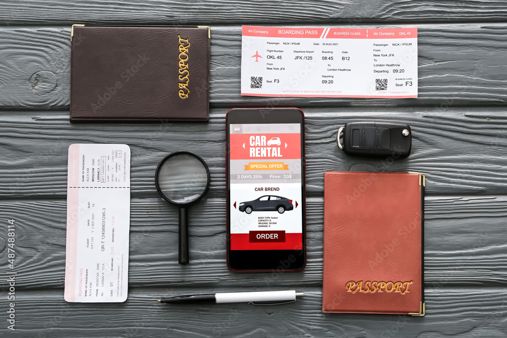 Mobile phone with open car rent app, passports, magnifier and tickets on dark wooden background