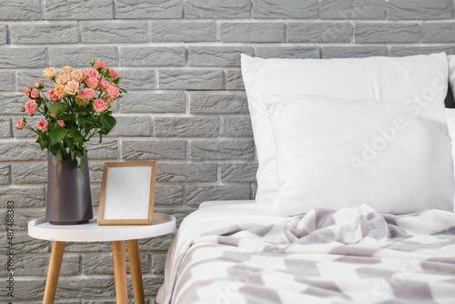 Vase with bouquet of beautiful fresh roses on bedside table near grey brick wall