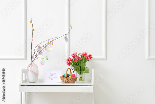 Beautiful Easter composition with basket, painted eggs, flowers and greeting card on table near white wall © Pixel-Shot
