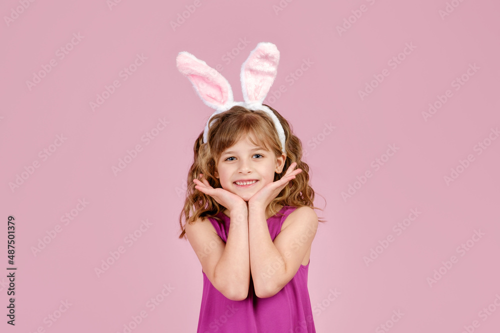 Adorable little coquette with long curly hair in stylish fuchsia colored dress and bunny ears touching cheeks and smiling at camera, against ink background in studio