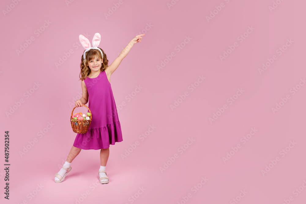 Full body little beautiful smiling girl in Easter bunny ears holds a basket with eggs on pink background and pointing away with hand. Copy space for text Full body little beautiful smiling girl in Easter bunny ears holds a basket with eggs on pink background and pointing away with hand. Copy space for text
