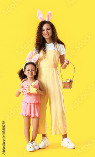 Little African-American girl with Easter rabbits, her mother and eggs on yellow background