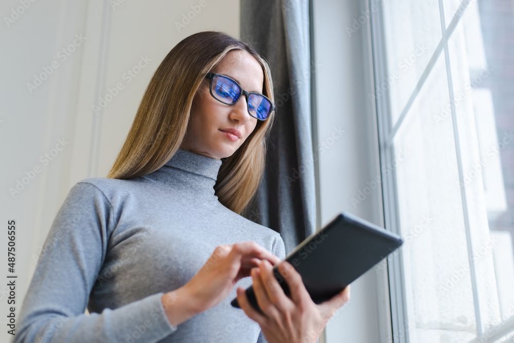 Young blonde woman browsing internet on tablet computer device at home.  Beautiful white female wearing nerd glasses and grey turtleneck shirt using  modern gadget for online communication Stock Photo