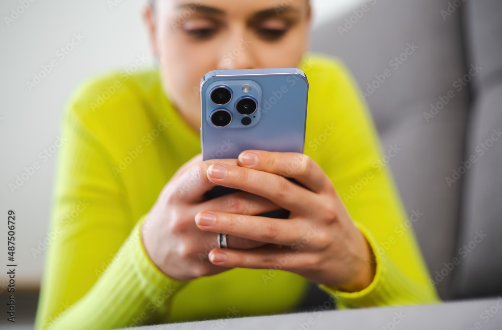 Young woman typing message on mobile phone at home while lying on couch. White female using modern smartphone for online communication during lockdown. Tomboy person texting with cellphone Young woman typing message on mobile phone at home while lying on couch. White female using modern smartphone for online communication during lockdown. Tomboy person texting with cellphone