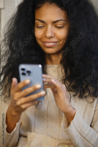 Happy black woman browsing internet on mobile phone. Cheerful African female using modern blue smartphone with triple camera. Young adult person communicating online with new cellphone gadget