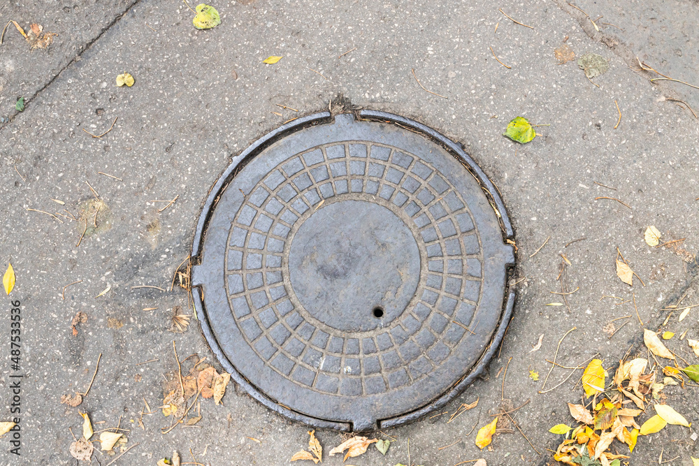 Old metal manhole cover sewer cap on the city street with yellow fall leaves