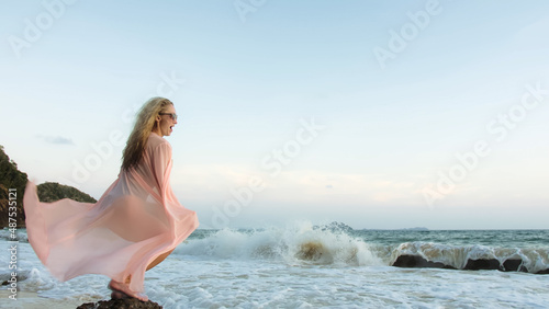 Attractive woman stand on a reef rock stone in sea on golden sunset. Girl on tropical beach in green swimsuit and flutter in wind pink tunic silk shirt cape  drinks her orange cocktail Pina Colada