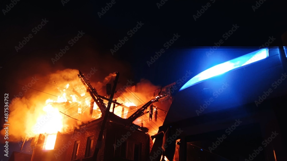 Fototapeta premium The roof of the house is on fire. The residential building burn, village. Firefighters put out a fire. Fire Department putting out flame. Fire truck, headlights and panel, siren, signal, alarm.