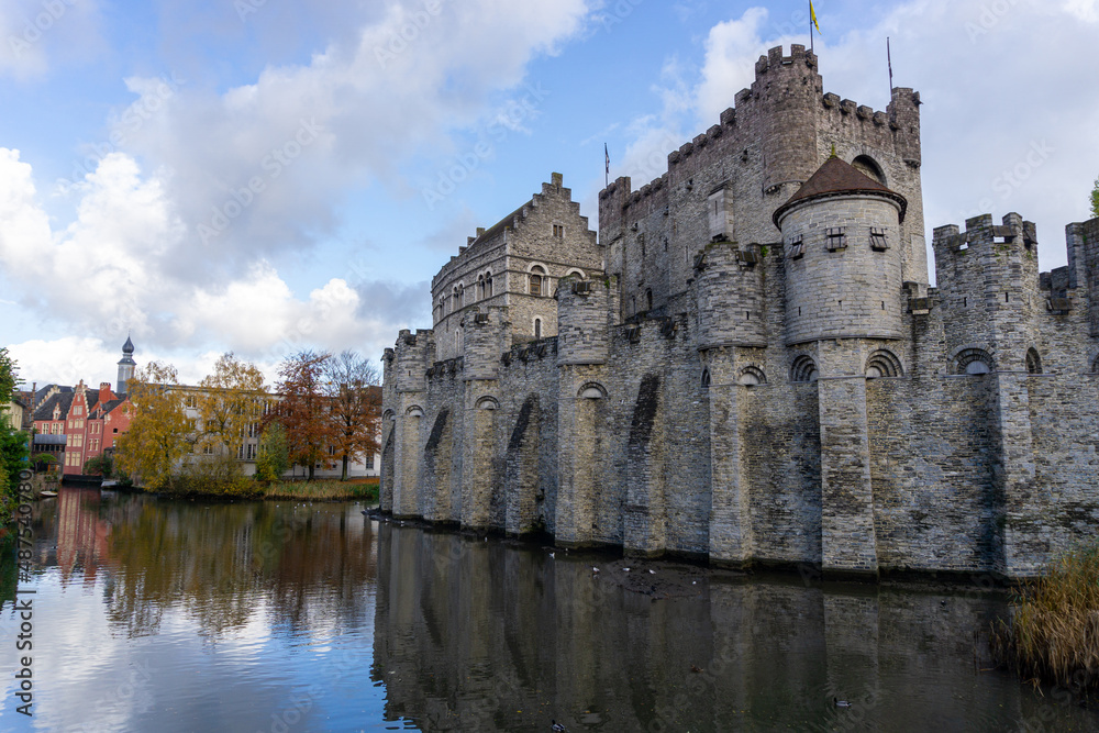 Ghent Castle with its reflection in the water on a day with some clouds. Trees at the end of the photo with fall colors.