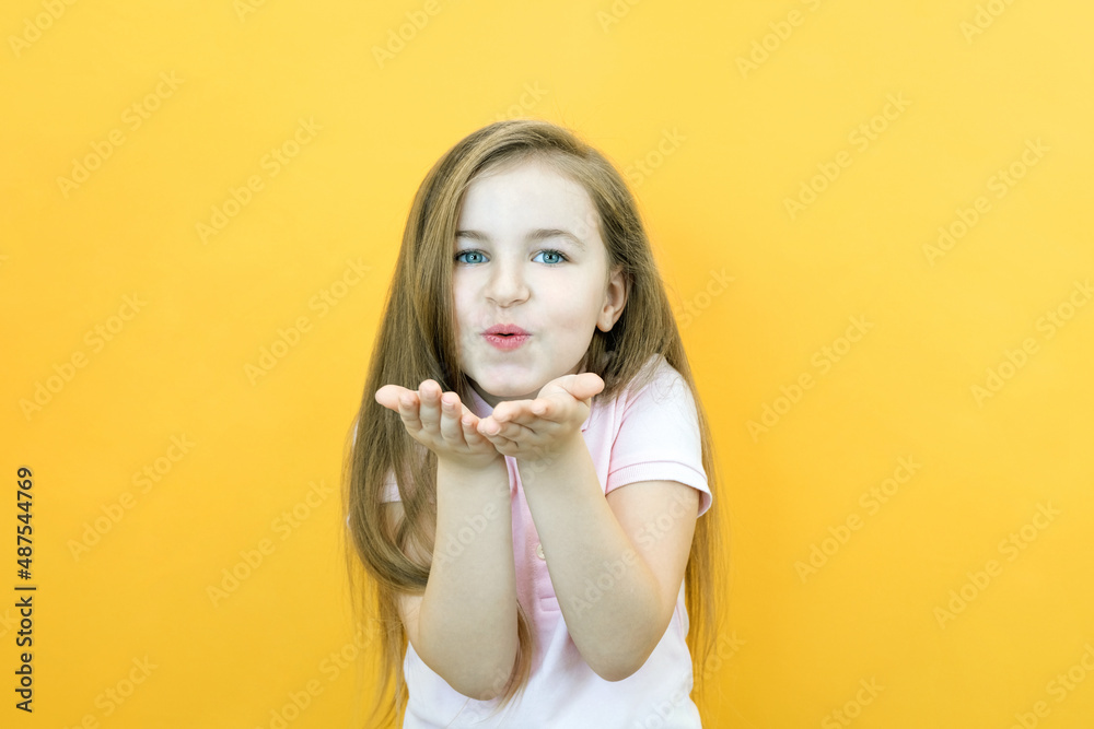 Little cute girl blows an air kiss directly to the camera on a yellow Little cute girl blows an air kiss directly to the camera on a yellow