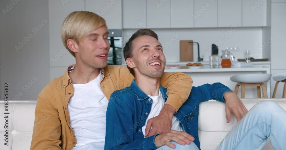 Happy men homosexual couple sit at home together on their sofa in an embrace. Male gay couple spend time at home. LGBT Relationship