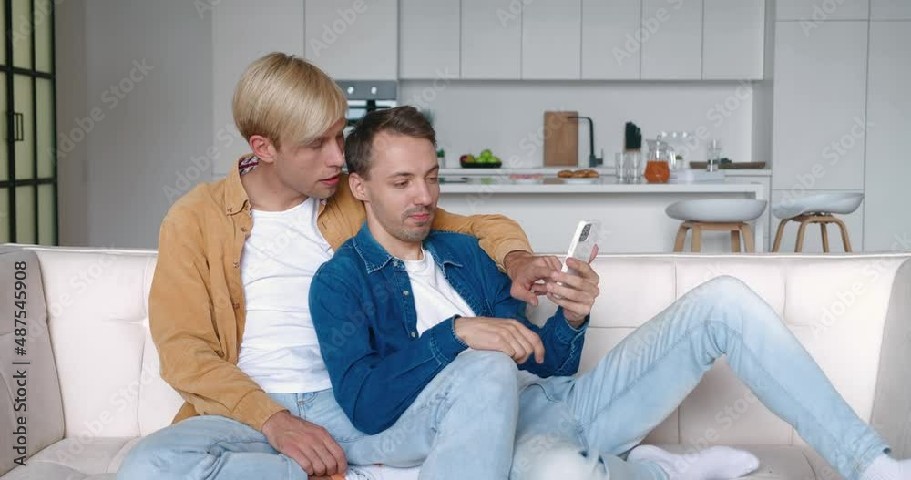 Happy men homosexual couple sit at home together in an embrace on their sofa and using smartphone. Male gay couple spend time at home. LGBT Relationship Happy men homosexual couple sit at home together in an embrace on their sofa and using smartphone. Male gay couple spend time at home. LGBT Relationship