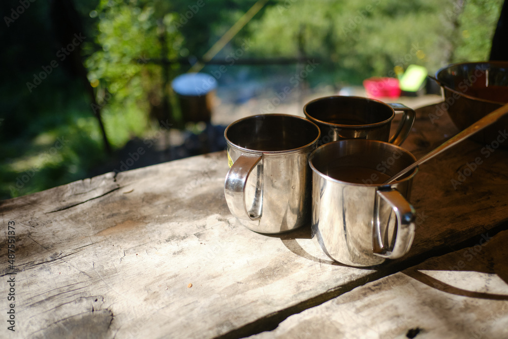 Three tourist iron mugs on the wooden table in a forest camping. Survival equipment for hiking concept. Close up photo of the 3 metal cup illuminated by the sun on timber desk. Journey, Trekking Theme Three tourist iron mugs on the wooden table in a forest camping. Survival equipment for hiking concept. Close up photo of the 3 metal cup illuminated by the sun on timber desk. Journey, Trekking Theme