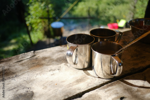 Three tourist iron mugs on the wooden table in a forest camping. Survival equipment for hiking concept. Close up photo of the 3 metal cup illuminated by the sun on timber desk. Journey, Trekking Theme