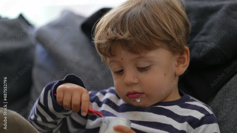 Child eating yogurt sitting on sofa one little boy eats yogurt with spoon Child eating yogurt sitting on sofa one little boy eats yogurt with spoon