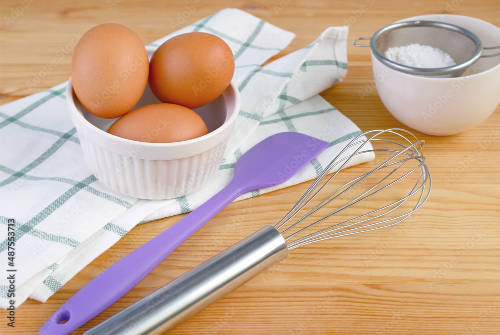 A whisk and spatula with eggs and kitchen utensils on wooden table A whisk and spatula with eggs and kitchen utensils on wooden table