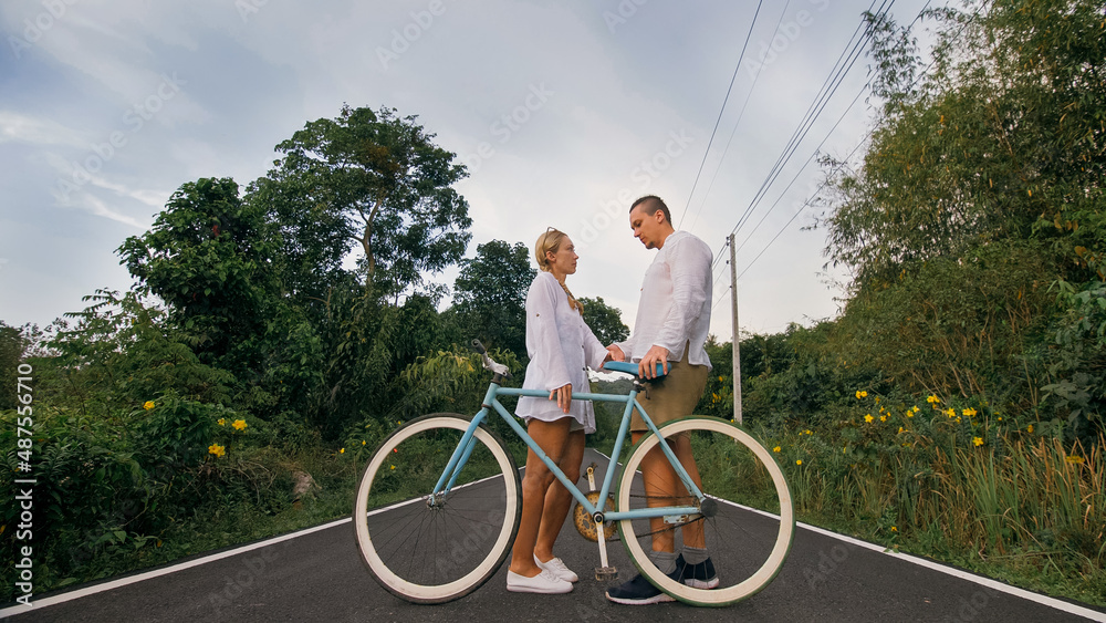 Obraz premium Biking road trip. Love couple on blue bike in white clothes on forest road. Just married woman and man kiss, hugs, stand on bicycle. Wedding, honeymoon. Cycling Cycle Fix. Asia Thailand ride tourism.