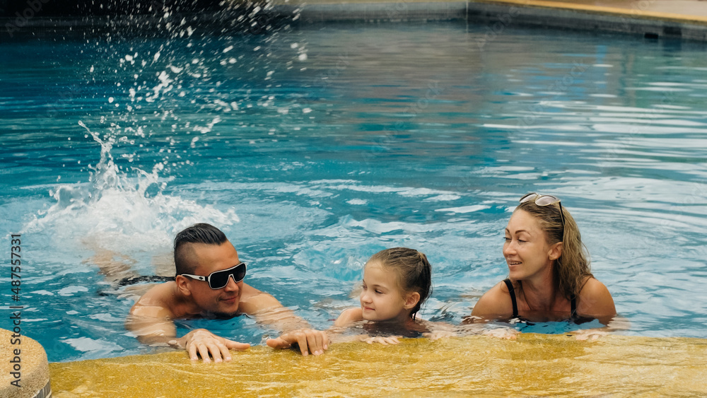 The mother and father with little daughter have fun in the pool. Mom and dad plays with the child. The family enjoy summer vacation in a swimming pool jumping, spinning, splash water. Slow motion.
