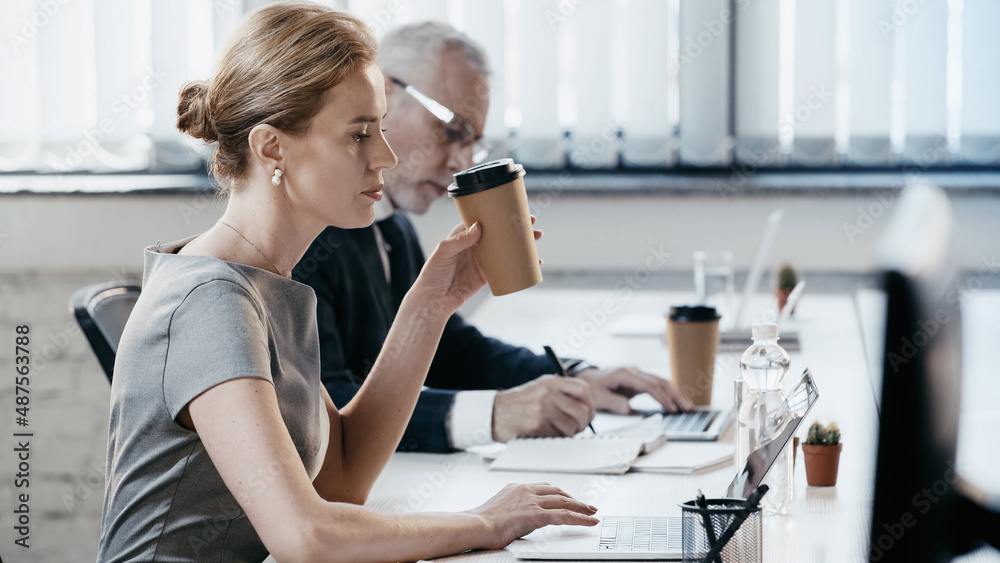 Side view of businesswoman holding paper cup and using laptop in office Side view of businesswoman holding paper cup and using laptop in office